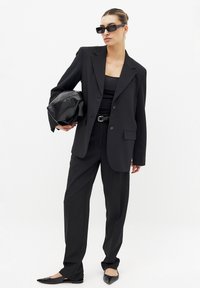 Costume noir ajusté avec une veste à un bouton, un pantalon large et un haut moulant. Les accessoires comprennent un sac à main noir et des lunettes de soleil.