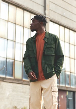 Chaqueta verde, ligera, con múltiples bolsillos, llevada sobre una camisa naranja y pantalones cargo beige, de pie frente a una gran ventana de cristal.