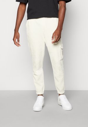 Cremfarbene Corduroy-Jogginghosen mit elastischen Bündchen und Seitentaschen, kombiniert mit weißen Sneakers. Das Oberteil ist ein schwarzes Kurzarmshirt.