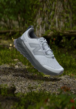 Pantof de alergare New Balance gri deschis, cu talpă groasă gri plutind deasupra unui drum pietros în mediul natural în aer liber.