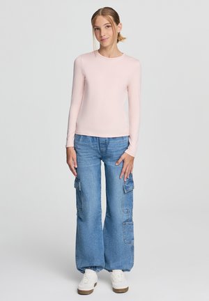 Top a maniche lunghe rosa chiaro con scollo rotondo abbinato a jeans blu a gamba larga con tasche cargo e una vestibilità rilassata. Sneakers bianche.