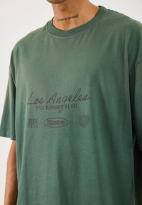 Mand med grøn t-shirt med teksten "Los Angeles 5102 Sunset Blvd," "1996," "turbo," og globus-logo tryk, plus "believe in yourself" tatovering på halsen.