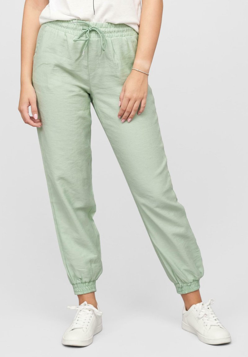 Pantalon de jogging vert clair avec taille élastique, à fermeture à cordon, poches et bas de jambes fuselés. Porté avec des baskets blanches.