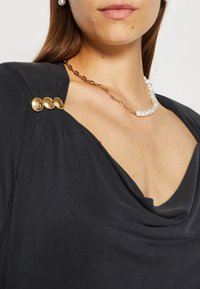 Top nero con scollo drappeggiato; presenta tre bottoni dorati e una collana in perle e oro. Tessuto liscio, design contemporaneo.