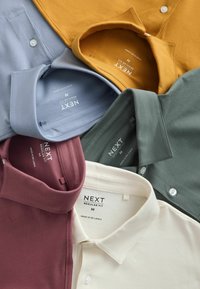 Cinq polos pliés de taille moyenne à coupe régulière en moutarde, bleu clair, vert, bordeaux et blanc, disposés en chevauchement sur une surface.