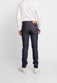 Tummansiniset farkut, kapealla istuvuudella, joissa on takataskut ja vyötäröllä vaaleanruskea nahkalappu. Yhdistetty valkoiseen pitkähihnaiseen paitaan.