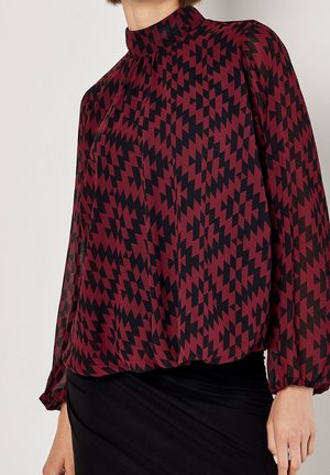 Frau trägt eine langärmelige bordeauxrote Bluse mit schwarzem geometrischem Zickzackmuster und einen schwarzen Rock, dargestellt von den Schultern bis zur Hüfte.