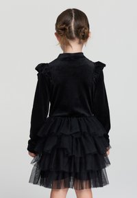 Abito nero con top in velluto e spalle arricciate, accompagnato da una gonna in tulle a strati che mette in risalto una silhouette ampia e un tessuto strutturato.