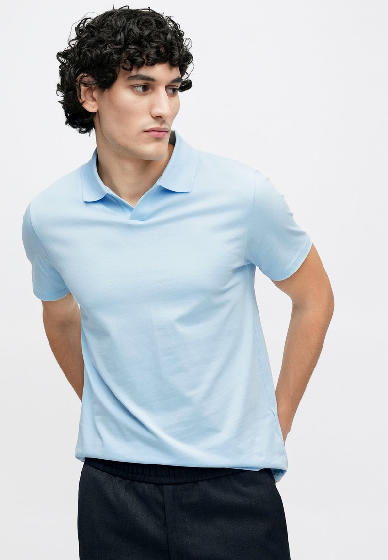 Hellblaues Polohemd aus glattem Baumwollstoff mit klassischem Kragen und kurzen Ärmeln, kombiniert mit dunklen Hosen mit elastischem Bund.