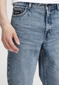 Gros plan d'une main reposant près de la poche d'un jean en denim bleu clair avec une étiquette de marque noire sur la petite poche.