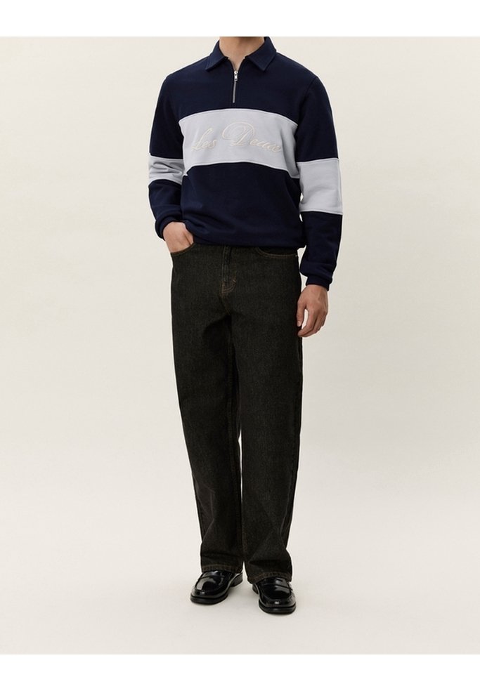 Homme portant un pull à col zippé à manches longues bleu marine foncé et gris, pantalon noir ample et chaussures noires, debout avec une main dans la poche.