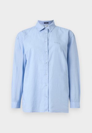 Chemise bleu clair à manches longues, boutonnée avec col et logo brodé discret sur la poitrine gauche, sur un fond uni.