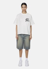 T-shirt bianca a maniche corte con scritta nera "Good for Nothing"; abbinata a pantaloni corti di jeans blu chiaro e sneakers bianche. Tessuto morbido.