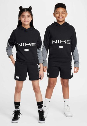 Zwei Kinder tragen passende schwarze Nike-Hoodies mit grauen Ärmeln, schwarze Shorts und sportliche Schuhe. Die Hoodies haben große weiße Markenschriften und Logos.