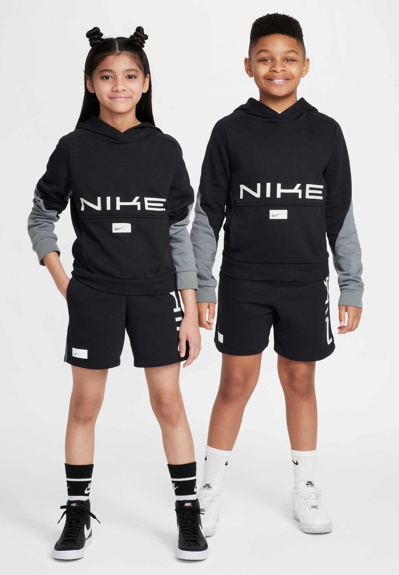 Två barn som bär matchande svarta Nike-hoodies med grå ärmar, svarta shorts och sportiga skor. Hoodies har stora vita varumärken och logotyper.