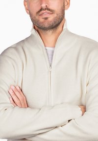 Suéter cremado con cremallera, con un patrón texturizado, cuello alto y mangas largas. La tela parece suave y ligera.