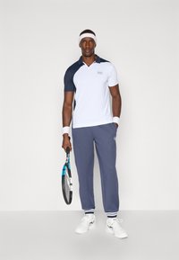 Tennis outfit featuring a white and navy short-sleeve polo with a logo, paired with navy sweatpants and white tennis shoes.