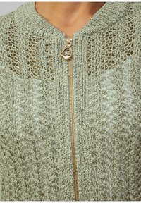 Jasnozielona sweterowa kurtka z otwartym splotem, z złotym zamkiem i teksturowanymi akcentami wokół kołnierza.