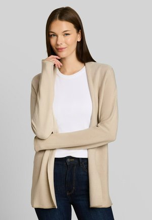 Junge Frau mit langen braunen Haaren, die eine beige offene Strickjacke über einem weißen Oberteil und dunklen Jeans trägt, lächelnd mit der Hand nahe am Kinn.