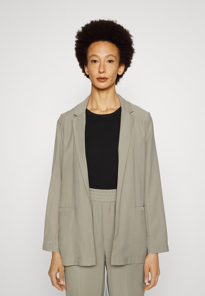 Blazer cinza claro com uma textura suave, apresentando um corte descontraído, lapelas recortadas e bolsos laterais, usado sobre uma blusa preta e calças a condizer.