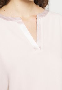 Helle rosa Bluse mit satinierter V-Ausschnitt, die über eine glatte Textur und ein klares Design verfügt. Der Ausschnitt hat einen dezenten Glanz.
