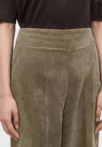 Personne portant un pantalon large en velours côtelé taille haute de couleur vert olive et une chemise à manches courtes marron foncé, mains détendues sur les côtés.