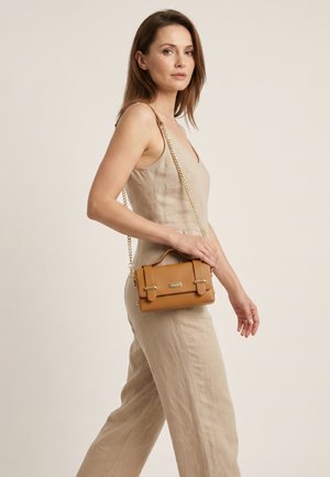 Vrouw in beige mouwloze jumpsuit draagt een kleine lichtbruine leren handtas met gouden kettingband over haar schouder, lopend tegen een effen achtergrond.