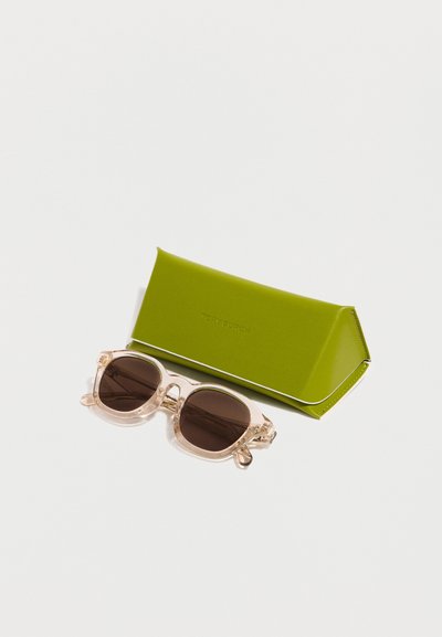 Γυαλιά ηλίου με διαφανή πλαίσιο και σκουρόχρωμους φακούς, τοποθετημένα δίπλα σε ένα ελαιόχρωμο τριγωνικό θήκη γυαλιών πάνω σε λευκή επιφάνεια.