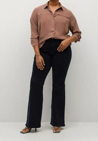 Model ma na sobie jasnobrązową koszulę z guzikami, założoną na ciemnoniebieskie dżinsy w stylu dzwonów, do których dobrała czarne sandaly na obcasie z odkrytymi palcami.