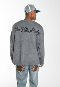 Grauer Strickpullover mit "Love Kills Slowly" in schwarzer Schreibschrift auf der Rückseite. Kombiniert mit hellblauen Jeans, die bunte Taschenmuster aufweisen.