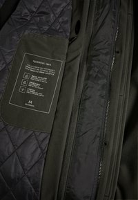 Interior de una chaqueta negra acolchada con una etiqueta de bolsillo verde que muestra información técnica. Características incluyen materiales repelentes al agua, transpirables y elásticos.