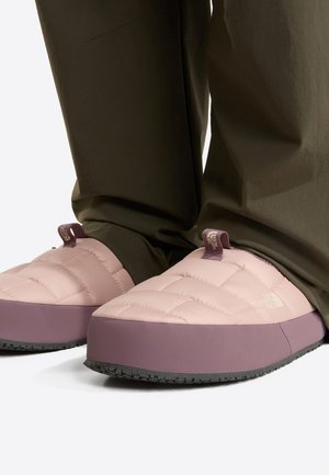 Zapatillas de estar por casa de color rosa pálido y malva, acolchadas, sin talón y con suelas negras, usadas con pantalones de color oliva oscuro.