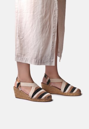 Sandale na platformu s klinastom petom u bež, crnoj i smeđoj prugastoj kombinaciji, s elastičnim remenčićima oko gležnja, nošene uz svijetlo bež suknju s bočnim prorezom.