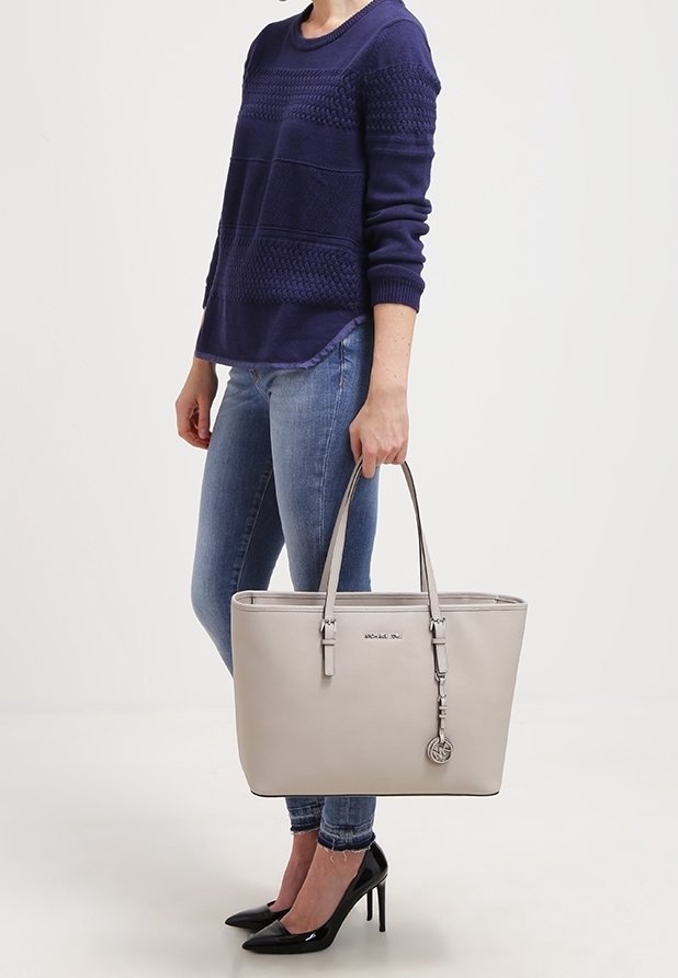 Sac fourre-tout gris clair avec poignées, design rectangulaire élégant, texture lisse, accent de logo et un charme attaché. Porté avec un pull bleu marine et un jean.