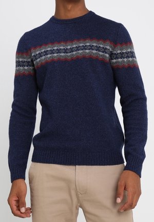 Mann trägt einen dunkelblauen Pullover mit roten, grauen und dunkelblauen Zickzack- und Sternmustern über der Brust und beige Hose.