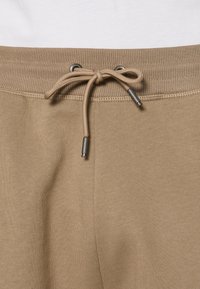 Pantalon de survêtement beige avec taille élastique côtelée et cordon de serrage noué avec embouts métalliques, présenté en vue rapprochée.
