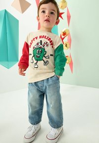 Crème shirt met lange mouwen met rode en groene mouwen, voorzien van een cartoonfiguur en tekst. Draag het met versleten blauwe jeans en witte sneakers.