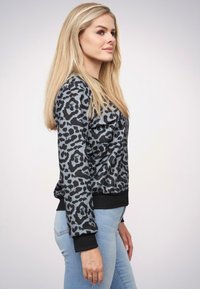 Blondynka zwrócona w prawo, ubrana w czarną i szaro-leopardzową bluzkę z długim rękawem oraz jasnoniebieskie dżinsy na jednolitym tle.