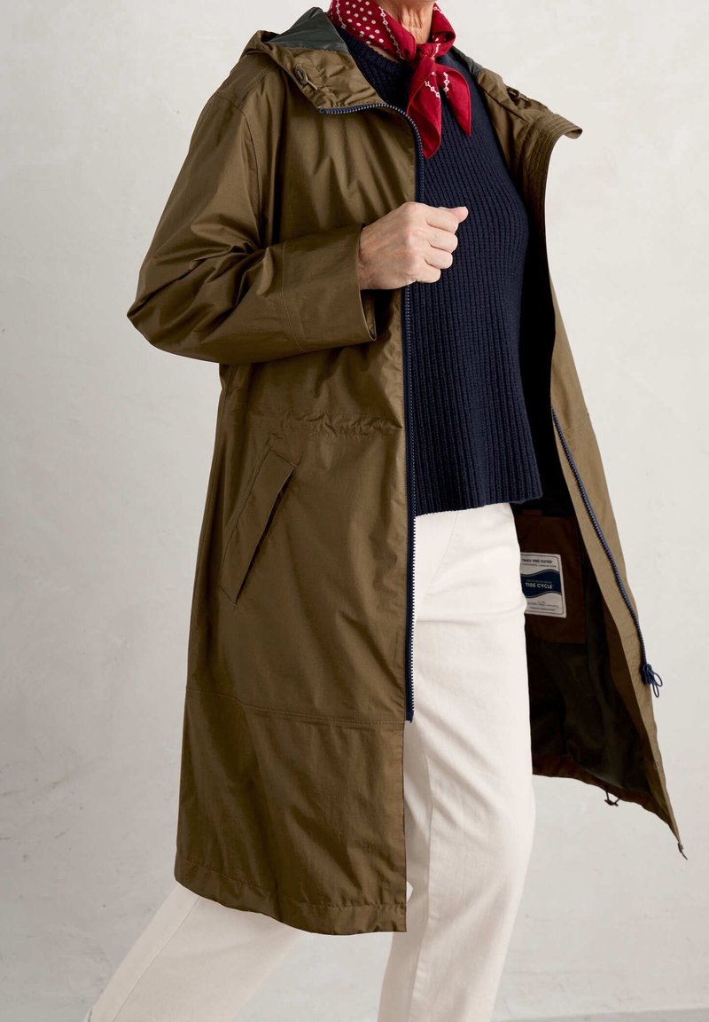 Osoba w oliwkowym płaszczu z kapturem, nosząca granatowy sweter, białe spodnie i czerwoną szalik w białe kropki, stoi na tle białego tła.