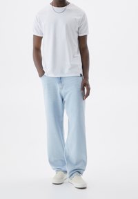 Hombre vestido con una camiseta blanca sencilla, jeans de pierna ancha azul claro, zapatos beige sin cordones y una cadena de plata, de pie con una mano en el bolsillo.