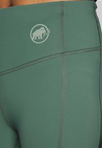 Leggings de sport verts en tissu extensible, avec des coutures à plat et un logo d'éléphant circulaire imprimé en gris clair.