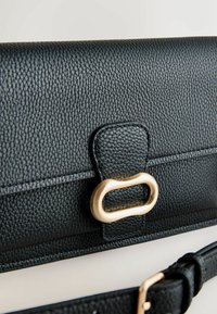 Črni usnjeni denarnica s teksturirano površino, opremljena z zlatim zaponko v obliki zanke. Vključuje odstranljiv pašček.