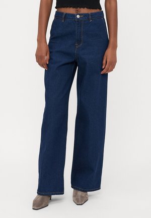 Person iført højtaljede, mørkeblå vidde jeans kombineret med taupe spidse ankelstøvler og en sort cropped top.