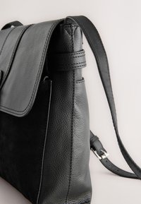Sac à bandoulière en cuir noir avec rabat texturé et bandoulière réglable, présenté sur un fond clair uni.
