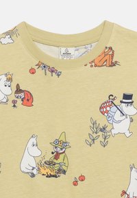 Ανοιχτό κίτρινο πουκάμισο με χαρακτήρες Μούμεν, συμπεριλαμβανομένων του Σνούφκιν που μαγειρεύει στη φωτιά και του Μούμεντρολ που κρατά μια τσάντα φορώντας καπέλο με κορδέλα.