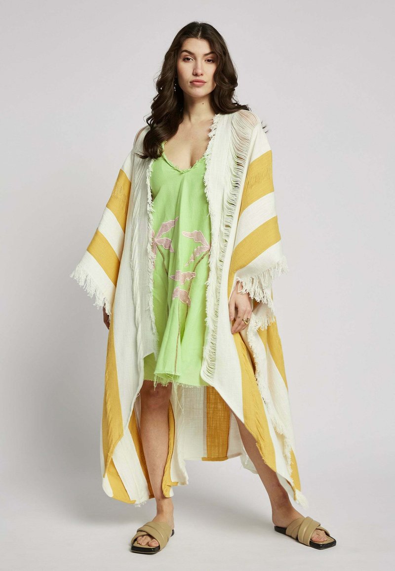 Lange kimono met witte en gele strepen en franjes, gelaagd over een groene jurk met een bloemenmotief, gecombineerd met beige schuiflats.