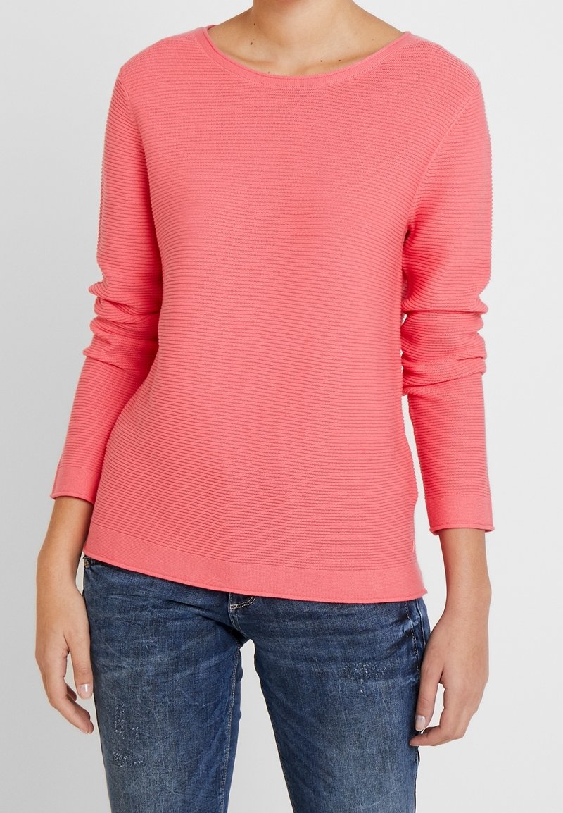 Różowa bluzka z długim rękawem o prążkowanej strukturze, z okrągłym dekoltem i podwiniętymi mankietami, zestawiona z niebieskimi dżinsami. Gładka tekstura, luźny fason.