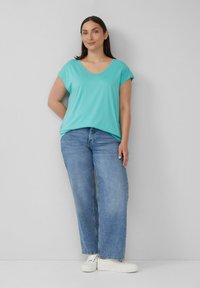 Lichtgroene korte mouwen V-hals T-shirt gecombineerd met lichtblauwe wijde jeans. Model draagt witte sneakers en staat tegen een effen achtergrond.