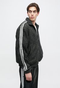 Μαύρο μπουφάν sherpa με ψηλό γιακά, λευκές ρίγες στα μανίκια, άνετη εφαρμογή και υφασμάτινη υφή. Το μοντέλο φοράει μαύρο παντελόνι.