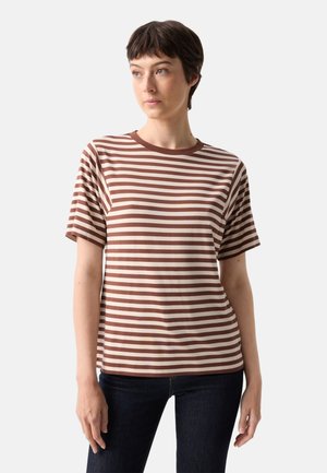 Kvinde med kort mørkt hår iført en brun og beige vandret stribet kortærmet skjorte og mørke bukser, stående foran en ensfarvet baggrund.